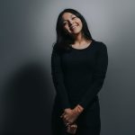 Simple portrait of a woman in black clothing smiling against a gray background.