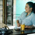 Side view of positive Asian female with dyed hair sitting at wooden counter with photo camera and refreshing drink in cafeteria